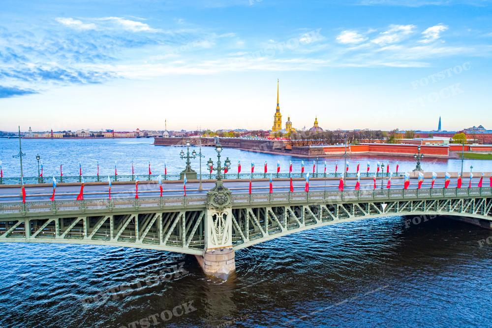 На фотографии изображен вид на Троицкий мост через реку Неву в Санкт-Петербурге. Мост соединяет Петроградскую сторону с центральной частью города. Вдалеке виднеется знаменитый шпиль Петропавловской крепости и золотые купола. Небо ясное с небольшими облаками, что придаёт фотографии живописность и простор. Это фото может использоваться для туристических путеводителей, исторических или культурных изданий, в статьях о путешествиях или как украшение в виде плаката или картины.